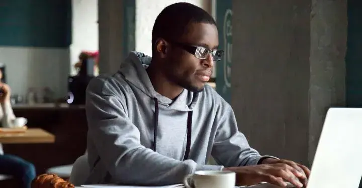 Man in gray sweatshirt types at laptop at desk with mug of coffee next to it