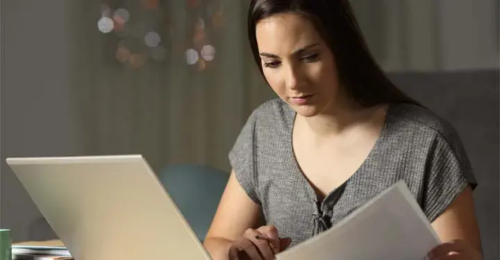 Woman sitting at laptop holding papers reviewing documents