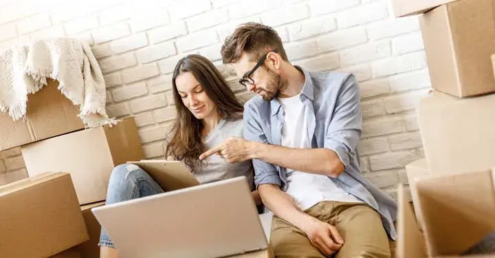 Relaxed man in glasses pointing to clipboard woman sitting beside him is holding surrounded by boxes