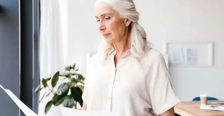 Older woman looking at paper while standing in front of window in room containing plant and table with coffee cup placed on it