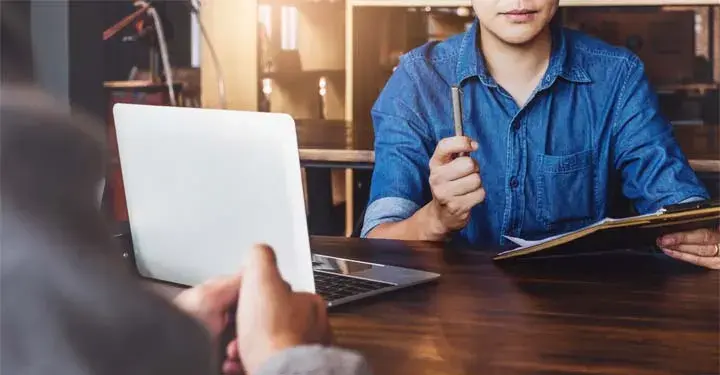 Person in denim shirt holding pen and clip board in front of computer with another person sitting across the table