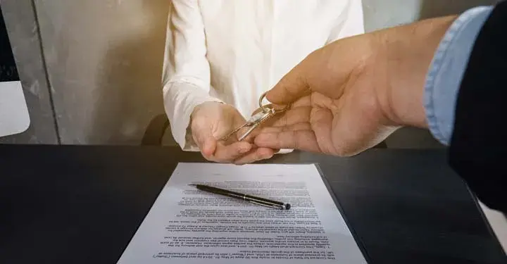 Hands passing house keys from one person to another.  Paperwork and a pen sit on the table