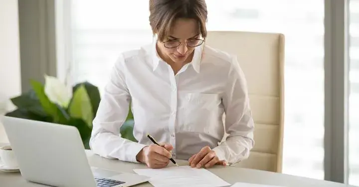 Woman in office holds pen above form at desk with laptop