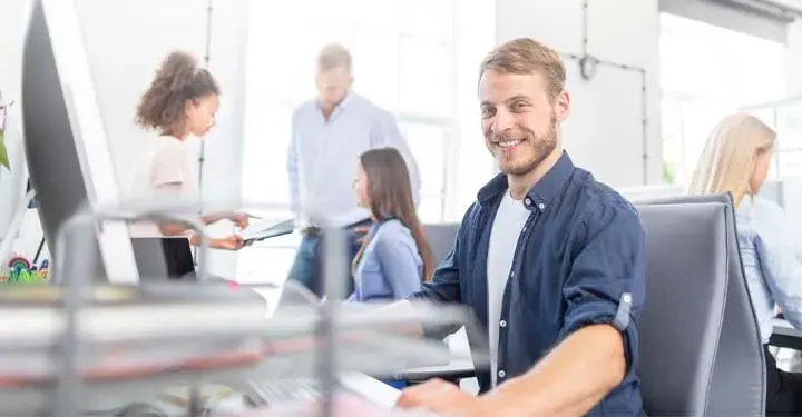 Bearded man smiles while at desk with other businesspeople behind him