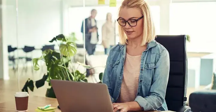 Woman in a pink shirt and denim jacket with glasses using her laptop in an office