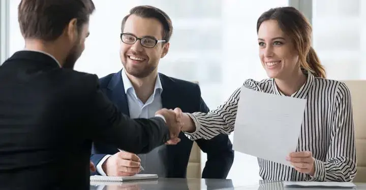 A smiling couple sitting across from a businessman.  The woman holds paperwork and shakes the businessman's hand.