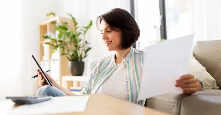 Woman in striped shirt and jeans sitting in living room and holding iPad and sheet of paper