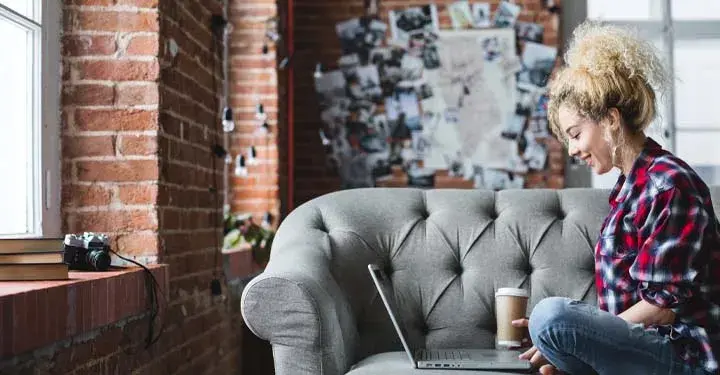 Smiling woman with a cup of coffee sitting on a couch looking at her computer