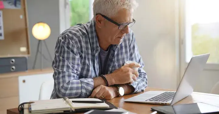 Man sitting at a table holding a cup of coffee looking at his laptop next to an open notebook