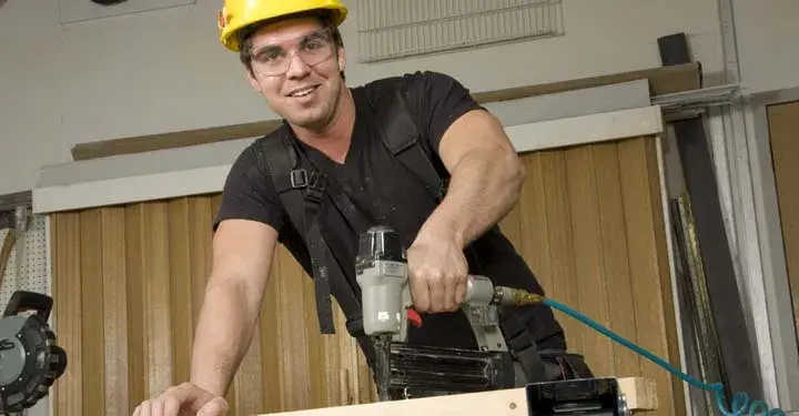 Construction worker in a hardhat and goggles holding a nail gun on a plank of wood