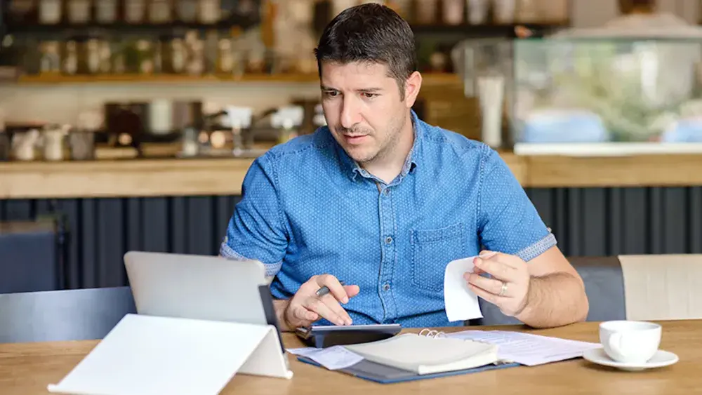 A cafe owner sits at a table looking at his tablet and using a calculator. Limited liability companies are popular among Florida entrepreneurs due to their flexible management structure.