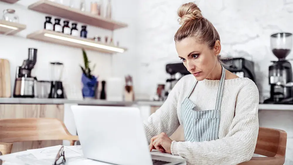 A woman who is opening a catering business reads her open laptop at her kitchen table. To open a business bank account, you'll need to obtain a certificate from the state, acquire an EIN, and provide the necessary documentation.