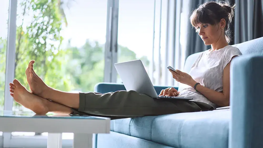 A woman seated on a couch with her bare feet on a coffee table checks her phone and her laptop after sending in her annual report. Submitting annual reports helps maintain your Florida LLC's compliance with state regulations and preserves its active status. 