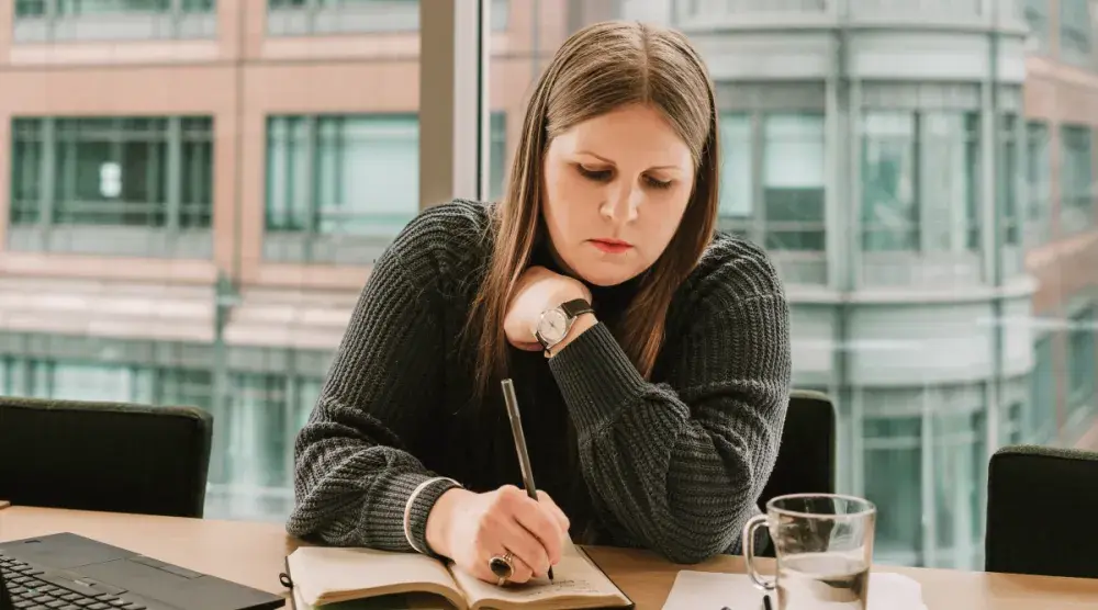 A woman sitting at a table in front of a laptop writing a draft of a demand letter in a notebook