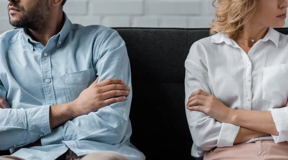 A couple considering divorce sits on a couch with their arms crossed, facing away from one another