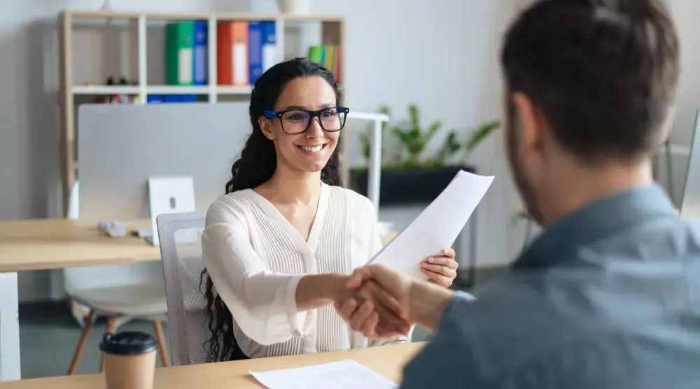 A woman sits at a desk holding an employment contract and nondisclosure agreement. She shakes hands with a man across the desk.