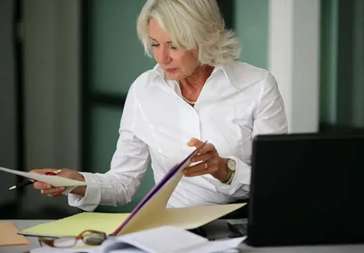 Businesswoman with grey hair holds paper and pen while looking through folders