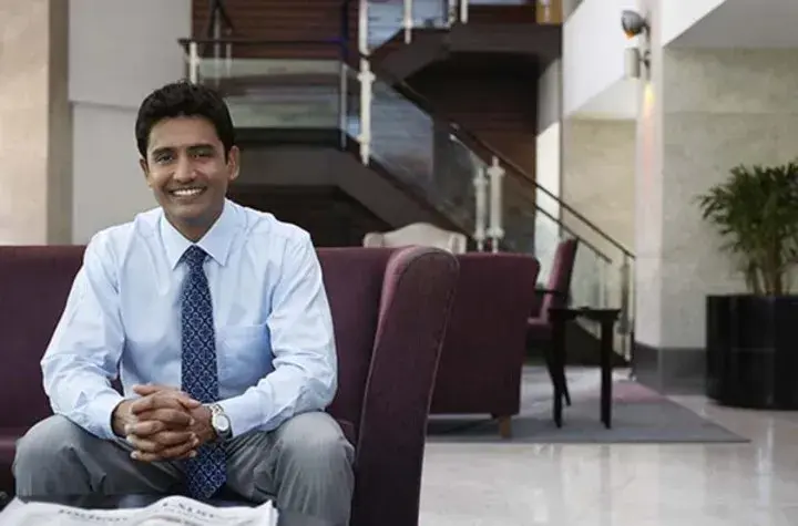 Man on a couch in a lobby smiling with a newspaper on the table in front of him