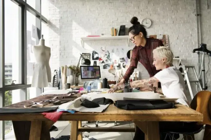 Two women, one seated in a chair and the other standing over her shoulder, work at a table covered in fabric samples