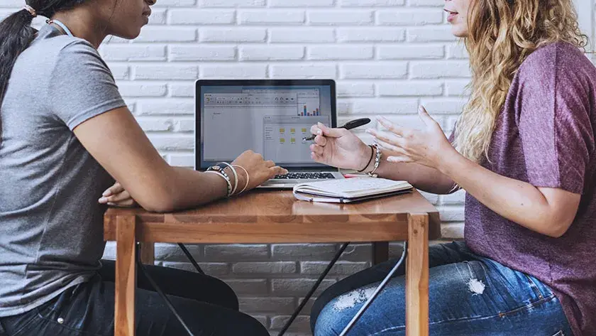 Two women talking side by side at table with laptop