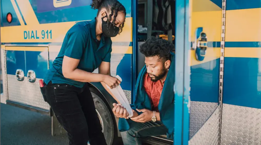 A woman wraps a white bandage on a man's hand as he sits on an ambulance step. Hiring a personal injury lawyer will help you recover damages if you are in an accident.