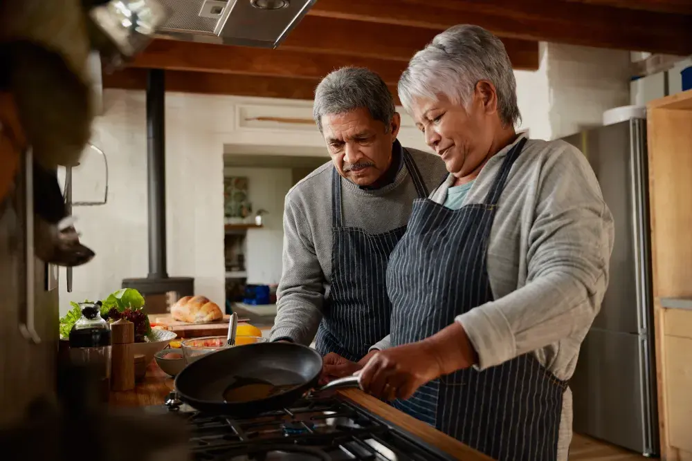 A man and woman, both wearing aprons, cook together on a stovetop in their kitchen.