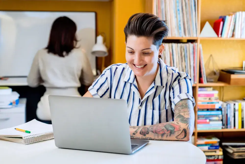 A woman with many colorful tattoos on her left arm sits at a table with an open laptop and notebook beside it as she applies for a legal name change in Texas.