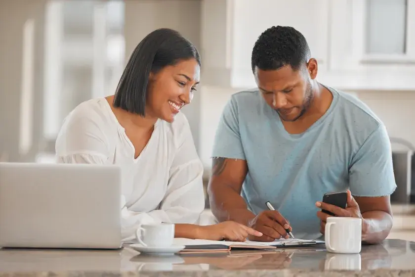 A woman seated at a table smiles as her fiance signs their prenup agreement.