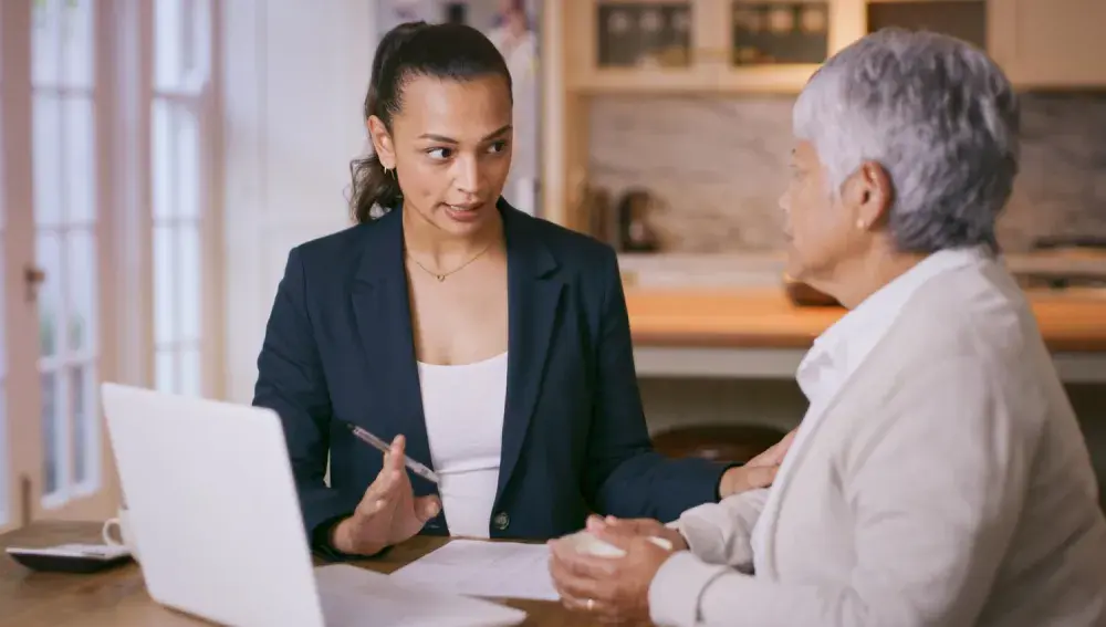 Two women, seated at a table with papers and an open laptop on it, discuss financial <a href=