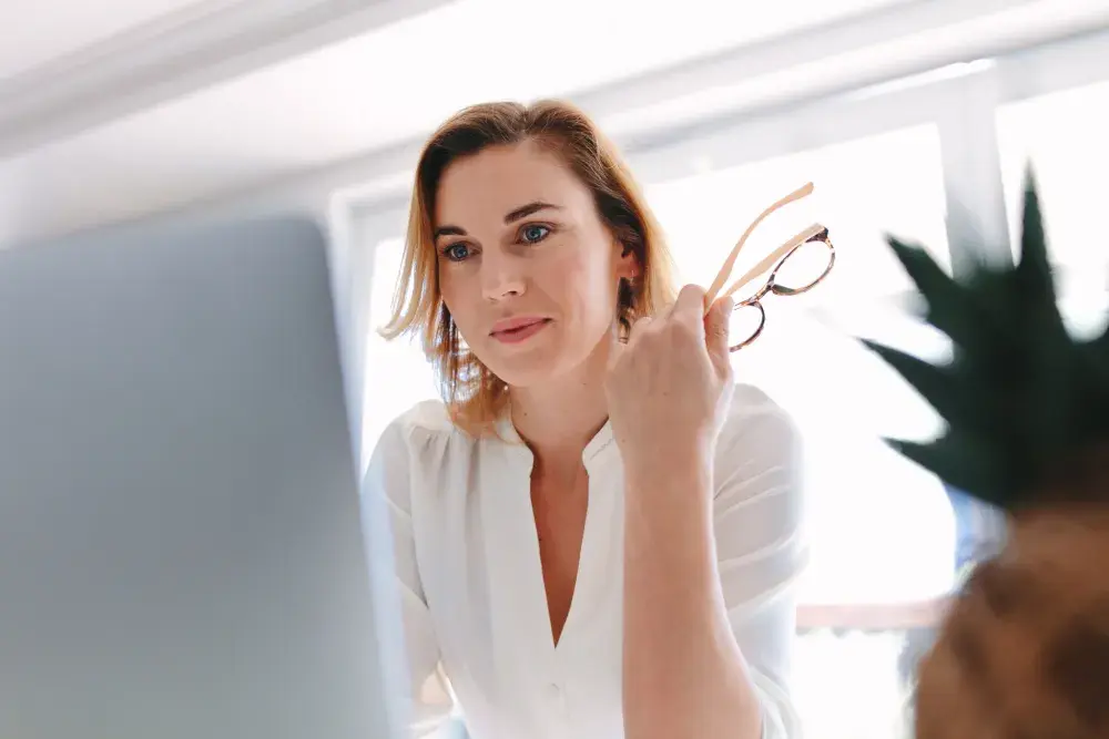 woman in white blouse holding glasses looking at her laptop