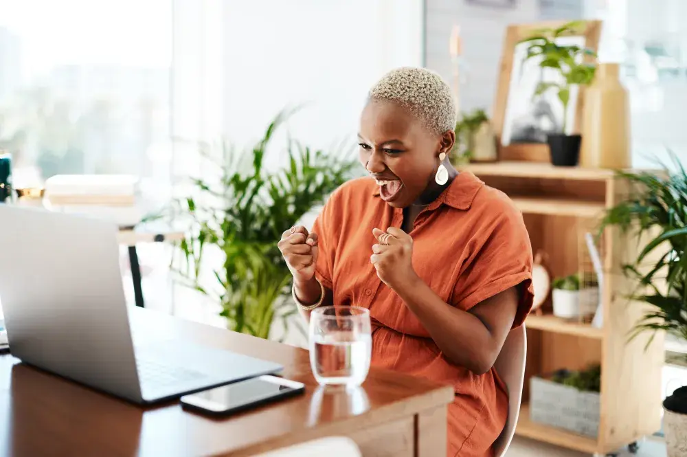 A woman seated at a desk with an open laptop pumps her fists after receiving notification her LLC was successfully formed.