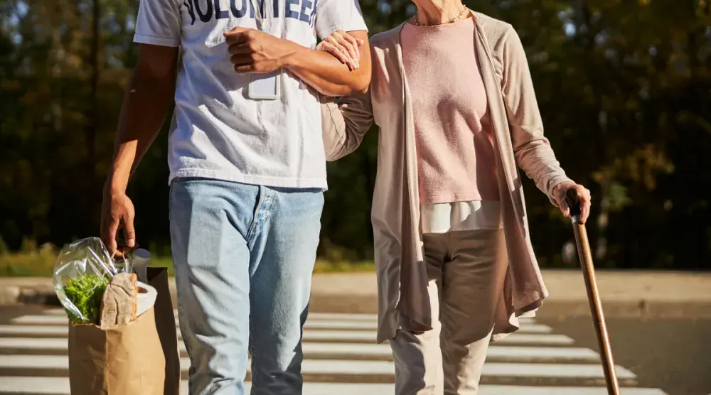 A man holding a grocery bag walking an older woman with a cane across a crosswalk to avoid a pedestrian accident