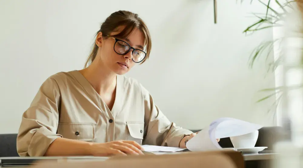 A woman wearing glasses sits at a desk and looks over prenup documents.